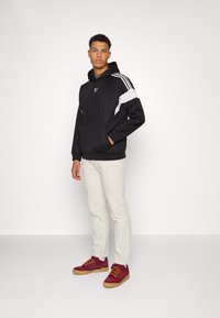 Felpa nera con cappuccio, pannello laterale bianco, tasca a marsupio e lacci. Abbinata a pantaloni beige chiaro e sneakers bordeaux.