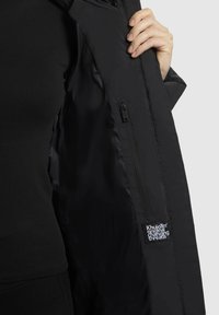 Zwarte jas binnenkant met een gladde textuur, voorzien van een ritszak en een label "Khujo Original Changing Threads."