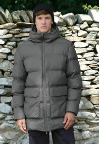 Geacă puf gri cu glugă, având secțiuni quilted, fermoar frontal și două buzunare mari în față. Modelul poartă o căciulă neagră.