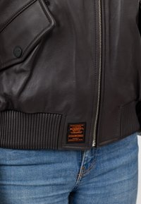 Brązowa skórzana kurtka bomberka z prążkowanymi mankietami i pasem, z przodem na zamek błyskawiczny i z logo na dolnym brzegu.