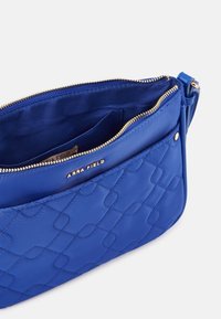 Borsa a tracolla sintetica blu trapuntata con hardware dorato, caratterizzata da una tasca esterna con zip e un interno in satin liscio. Etichetta del marchio visibile all'interno.