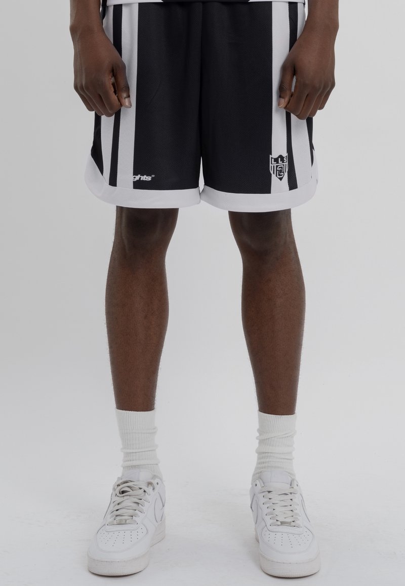 Personne portant un short de sport rayé noir et blanc, des chaussettes blanches hautes et des baskets blanches, debout devant un fond uni.