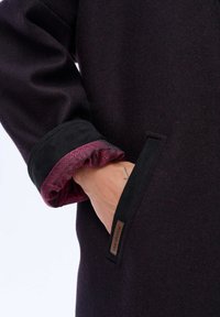 Zwarte en bordeauxrode jas van een textuurstof. Beschikt over een modellen binnenmanchet, zijzak en een merklabel aan de zoom.