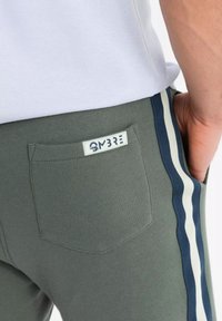 Pantalon de survêtement gris avec une poche arrière et une étiquette. Présente des bandes latérales en bleu marine et blanc. Fabriqué en tissu doux.