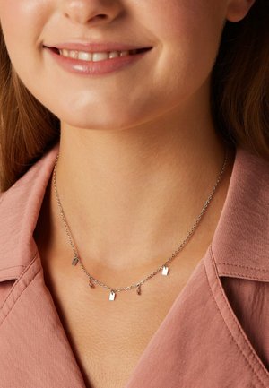 Vrouw draagt een delicate zilveren ketting met kleine vierkante en kralen bedeltjes, gekleed in een rozenkleurig overhemd met kraag.