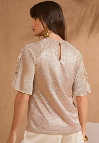 Top a maniche corte in tessuto rosa chiaro con riflesso metallico, caratterizzato da un dettaglio a goccia sulla schiena e delicati accenti arricciati sulle maniche.