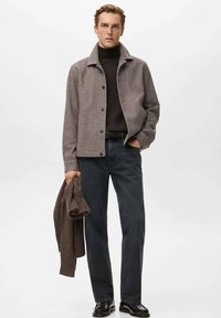 Chaqueta gris de mezcla de lana con cuello, usada sobre un suéter de cuello alto negro, combinada con jeans oscuros; sujetando un suéter de punto marrón. Zapatos negros.