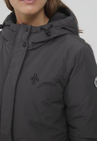 Mørkegrå polstret jakke med hette, lukkningsknapper og logo broderi på brystet, med jevn, lett gloss aktig tekstur.
