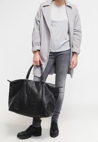 Schwarze Leder-Tote-Tasche mit strukturiertem Oberfläche und zwei Griffen, kombiniert mit grauer Kleidung, die einen Mantel und schmale Jeans umfasst.