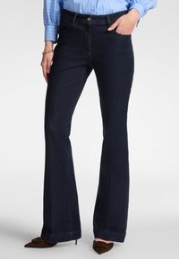 Tmavě modré zvonové džíny z denimové látky s hladkou texturou. Mají zapínání na přední knoflík a kapsy, stylizované s hnědými špičatými podpatky.