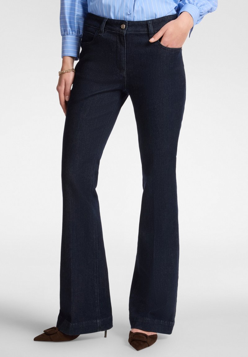 Tmavě modré zvonové džíny z denimové látky s hladkou texturou. Mají zapínání na přední knoflík a kapsy, stylizované s hnědými špičatými podpatky.