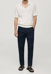 Polo blanc à manches courtes avec un tricot structuré et un col en V, associé à un pantalon bleu marine foncé et des sandales noires. Design simple et épuré.