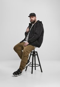 Svart zip-up hettegenser med en grå t-skjorte, brune cargo-bukser med lommer og svarte joggesko. Sitter på en svart krakk mot en hvit bakgrunn.