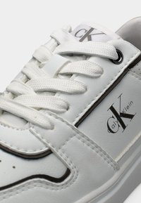 Άσπρο δερμάτινο αθλητικό παπούτσι με άσπρα κορδόνια, μαύρες λεπτομέρειες και λογότυπο στο πλάι. Το παπούτσι έχει λεία υφή με ραμμένες λεπτομέρειες.