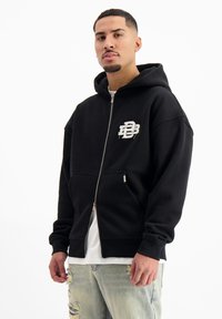Zwarte rits-hoodie met een voorste kangoeroezak, voorzien van een wit logo op de borst, gemaakt van fleecemateriaal met een zachte textuur.