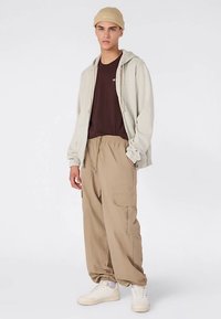 Jasnoszara bluza rozpinana na zamek, brązowy t-shirt, beżowe spodnie typu cargo i kremowe sneakersy. Model nosi beżową dzianą czapkę. Casualowy strój z różnorodnymi fakturami.