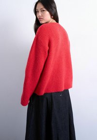 Jeune femme aux longs cheveux noirs portant un pull rouge vif texturé et une jupe foncée, regardant par-dessus son épaule sur un fond uni.