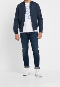 Veste bomber marine en matériau léger, t-shirt blanc avec graphique, jean bleu foncé et baskets blanches avec les ourlets retroussés.