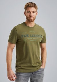 Olive zelené bavlnené tričko s krátkymi rukávmi a okrúhlym výstrihom. Obsahuje potlačený text vrátane "PME Legend" a letecké detaily.