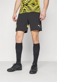 Svarta idrottsshorts med gula accenter, kombinerade med svarta strumpor och svarta sportskor med en strukturerad design.