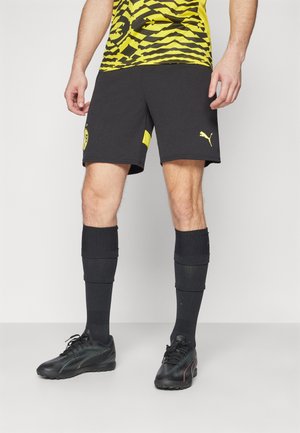 Schwarze Sportshorts mit gelben Akzenten, kombiniert mit schwarzen Socken und schwarzen Sportschuhen mit strukturiertem Design.