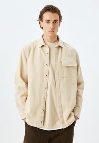Camisa de algodón beige con frente abotonado, mangas largas, bolsillo en el pecho y botones en tono dorado. Tejido ligero y texturizado.