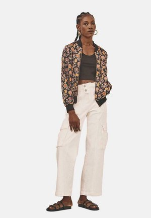 Chaqueta bomber con estampado floral en negro, top corto oscuro, pantalones cargo blancos y sandalias negras. La chaqueta presenta puños y dobladillo acanalados.