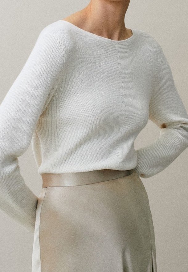 Femme portant un pull à manches longues côtelé blanc rentrée dans une jupe satinée beige, les mains posées sur les hanches devant un fond neutre.