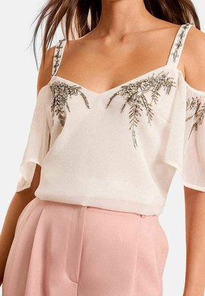 Donna che indossa un top bianco con spalle scoperte e decorazioni di perline d’argento e pantaloni rosa chiaro a vita alta, mostrata dal collo ai fianchi.