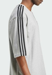 T-shirt grigia a maniche corte con design a tre strisce nere sulle spalle, realizzata in morbido tessuto di cotone, con scollatura a girocollo.