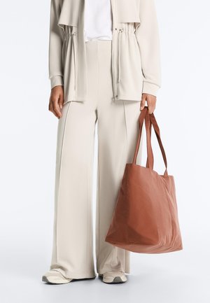 Personne portant un pantalon beige à jambes larges, une veste assortie, des baskets blanches, tenant un grand sac fourre-tout marron à côté.