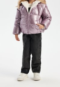 Enfant portant une doudoune lavande brillante avec doublure en polaire blanche, un jean noir et des baskets blanches, se tenant devant un fond uni.