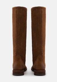 Kaksi korkeaa ruskeaa mokkasiinisaapasta, joissa on ommeltu X-kuvio varressa, litteät puukeng�ät ja pyöristetyt varpaat, nähtynä takaa.