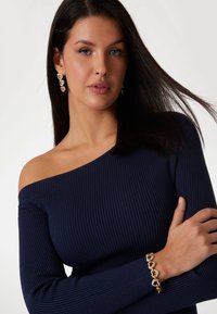 Maglione a coste blu navy con scollo a spalle scoperte e maniche lunghe, abbinato a un bracciale con motivo a cuori in oro. Texture liscia e design aderente.