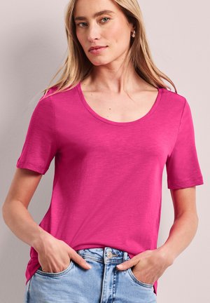 Femme aux longs cheveux blonds portant un t-shirt rose vif à manches courtes avec un col rond et un jean bleu clair, les mains dans les poches, sur un fond neutre.