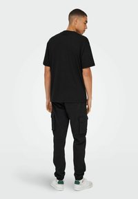 Camiseta negra de algodón combinada con pantalones cargo negros. Los pantalones cuentan con bolsillos laterales y puños elásticos, complementados por zapatillas blancas con acentos verdes.