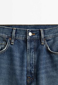 Gros plan de la ceinture d'un jean en denim bleu avec fermeture par bouton, passants de ceinture, poches avant et détails de couture visibles.