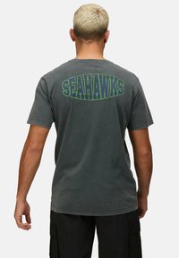 Graues kurzärmeliges T-Shirt mit der Aufschrift "SEAHAWKS" in fettem grünen Text auf der Rückseite. Aus weicher Baumwolle mit einem leicht verwaschenen Finish gefertigt.