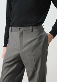 Persona con pantalones grises a la cintura, de corte sastre, con cierre de botón y cremallera, mano en el bolsillo, y una camiseta negra de manga larga.