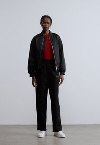 Czarna kurtka typu bomber na czerwonej koszuli, w połączeniu z czarnymi spodniami dresowymi i białymi trampkami. Gładka tkanina z minimalistycznym wzornictwem i funkcjonalnymi detalami.
