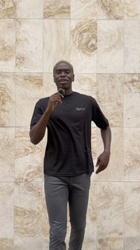 Svart kortermet skjorte med hvit logo, kombinert med grå bukser. Bakgrunnen har lyse steinfliser med et naturlig mønster.