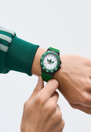 Zeleno uro z belim številčnicom, črnim ohišjem in zelenim okvirjem. Ima gumijast trak. Vidne kazalce in urne oznake. Prilagodljivo na zapestju.