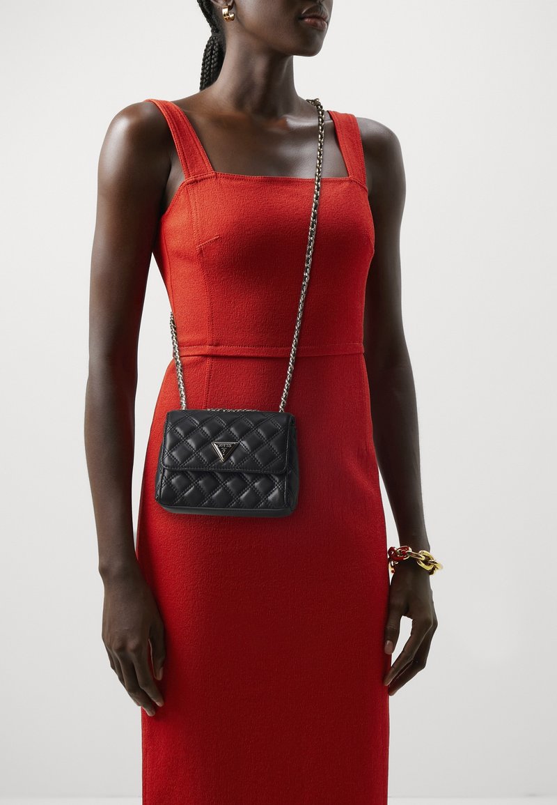 Kobieta ubrana w czerwoną sukienkę bez rękawów, z czarną pikowaną torebką na ramię i masywną złoto-czerwoną bransoletką na prawej ręce.