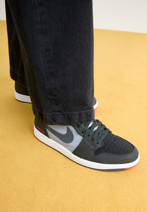Baskets Nike noires et grises avec semelles blanches et accents rouges portées avec un pantalon noir sur un sol jaune.