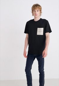 T-shirt noir avec une poche beige carrée au motif découpé. Le mannequin porte un jean foncé ajusté. Tissu lisse et design décontracté.