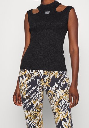 Ærmeløs sort ribstrikket top med udskæringer ved skuldrene, båret sammen med sorte bukser med hvid tekst og gyldne barokmønstre.