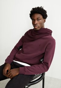 Sudadera marrón con un detalle de bufanda texturada, cuello redondo y dobladillo inferior blanco en contraste. Pantalones negros, sentado en una silla negra.