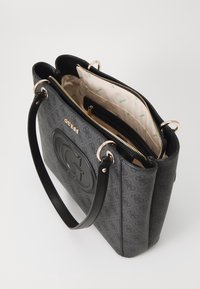 Harma ja must Guess-käekott, millel on reljeefne logo, kuldne riistvara, kaks kätet ja beež vooder koos lukuga sektsioonidega.