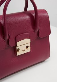 Furla-väska i djup vinröd, texturerad läder med lås i guldton, rundade handtag och en slank, strukturerad design.