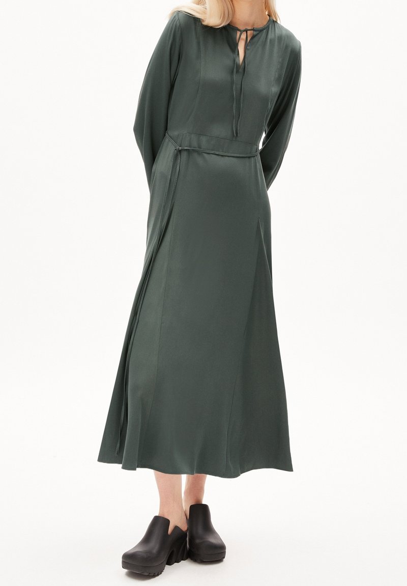 Pitkä, tummanvihreä satiininen mekko, jossa on avainreikäkaula-aukko, pitkät hihat ja liehuva helma. Yhdistetty mustiin massiivisiin puukenkään.